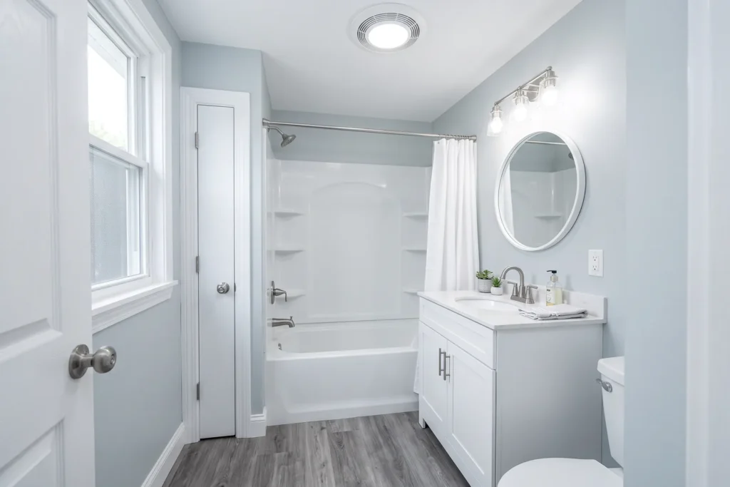 Refreshed small bathroom keeping its original layout, with a new acrylic tub and shower surround, white vanity, round mirror, and gray vinyl plank floor